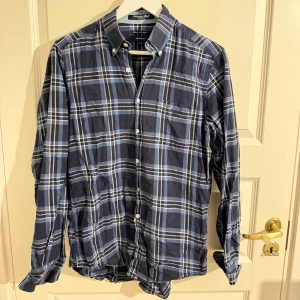 Blårutig skjorta från Gant - Snygg blårutig skjorta från Gant med klassisk button-down krage och vita knappar. Skjortan är tillverkad i bomull och har lång ärm. Perfekt för dig som gillar en tidlös och avslappnad stil.