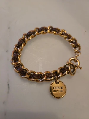 Guldfärgat armband från Guess - Snyggt giraffmönstrat armband från Guess med grova guldfärgade länkar och en rund berlock med texten 'GUESS EST. 1981'. Armbandet har en klassisk kedjedesign och stängs med en karbinhake. Perfekt accessoar för att lyfta din outfit.