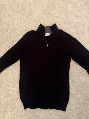 Svart halvzip-tröja John Henric - Klassisk svart stickad tröja från John Henric med halvzip och hög krage. Perfekt för lager på lager och enkel att matcha med olika outfits. Tröjan har långa ärmar och ribbade muddar vid ärmslut och nederkant.