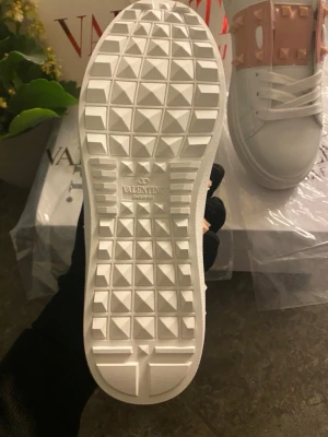 Valentino Rockstud vita sneakers - Säljer ett par vita Valentino Rockstud sneakers med rosa detaljer och ikoniska nitar. Skorna har snörning, platt sula och är tillverkade i skinn. Snygg och modern design med coola pyramidnitar på ovandelen.