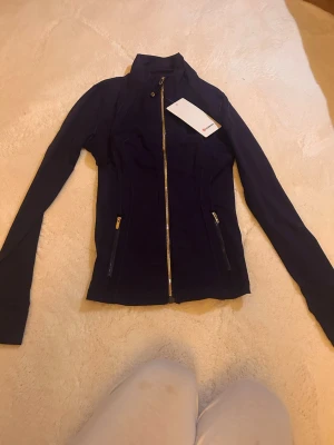 Marinblå träningsjacka från Lululemon - Slimmad marinblå träningsjacka från Lululemon med hel dragkedja i guld och två fickor med dragkedja framtill.  Storlek M men hade passat S