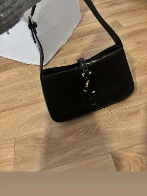 Svart handväska med gulddetaljer - Stilren svart handväska i skinn med justerbart axelband och guldfärgat spänne framtill. Väskan har en avlång, rektangulär form och stängs med dragkedja. Perfekt accessoar för att lyfta din outfit. Komtakta för fler bilder