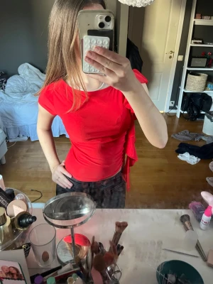 Röd topp med volang och knyt - Snygg röd topp med korta ärmar och volangdetalj på sidan samt knyt i midjan. Modellen är figurnära och toppen har en enkel, stilren design som passar perfekt till jeans eller kjol. Materialet känns mjukt och stretchigt.