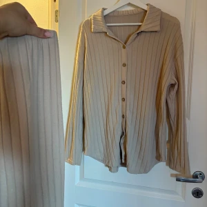 Beige ribbat loungewear-set Chiquease - Mysigt loungewear-set i beige ribbat material. Setet består av en långärmad skjorta med krage och knappar framtill samt matchande byxor med hög midja. Perfekt för chill dagar hemma eller när du vill vara bekvämt stylish.