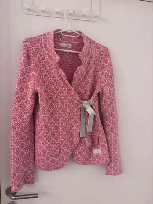 Mönstrad kofta från Odd Molly - Säljer en rosa och vit mönstrad kofta från Odd Molly med dekorativ knytning i sidenband på sidan. Koftan har långa ärmar, två fickor fram och en unik kant runt halsen. Perfekt för dig som gillar färg och detaljer!