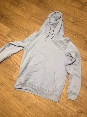 Ljusblå hoodie från OTP Leisurewear - Snygg ljusblå hoodie från OTP Leisurewear med klassisk känguruficka och dragsko i huvan. Perfekt för en avslappnad stil och enkel att matcha med jeans eller joggers. Mjuk bomullskänsla och ribbade muddar vid ärmslut och nederkant.