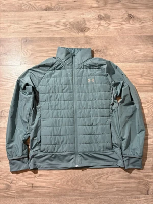 Under Armour - Snygg mintgrön quiltad jacka från Under Armour med dragkedja framtill och hög krage. Jackan har diskret logga på bröstet och är tillverkad i lätt polyester, perfekt för kyliga dagar. Passformen är normal och jackan har långa ärmar. Storlek L passar också M