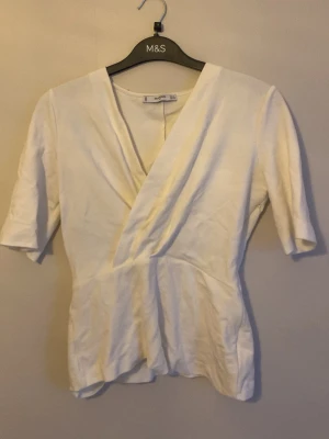 Vit omlottopp från mango - Säljer en stilren vit omlottopp från mango med v-ringning och halvlånga ärmar. Toppen har en figurnära passform och är tillverkad i mjuk bomull, perfekt för en clean och fräsch look. Enkel att matcha med både jeans och kjol.