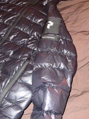 Svart dunjacka Peak Performance - Snygg svart dunjacka från Peak Performance med quiltad design och dragkedja framtill. Jackan har två sidofickor med dragkedja och en logga på ärmen. Perfekt för kalla dagar och enkel att matcha med olika outfits.