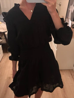 Neo noir klänning  - Svart kort klänning med v-ringning, långa ärmar och volangdetaljer vid axlar och nederkant. Klänningen har markerad midja och är tillverkad i ett lätt tyg som ger en luftig känsla. Perfekt för dig som gillar en stilren och trendig look. Aldrig använd 💕