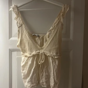 Beige stickad linnetopp med volang - Supersöt beige linnetopp med stickad överdel, volangdetaljer på axelbanden och knyt framtill. Toppen har djup v-ringning och är ärmlös, perfekt för varma dagar. Ger en romantisk och bohemisk känsla till din outfit.