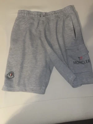 Grå Moncler shorts med ficka - Snygga grå shorts från Moncler med elastisk midja och dragsko. Stora sidofickor, varav en med lock och Moncler-logga broderad på benet. Perfekta för sommaren och chill dagar.