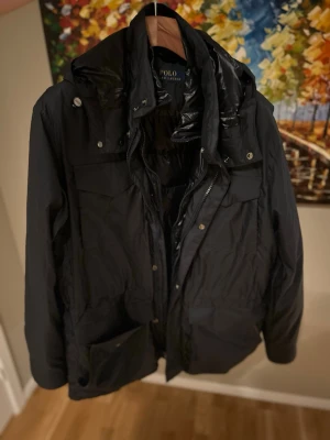 Svart dunjacka från Polo Ralph Lauren - Stilren svart dunjacka från Polo Ralph Lauren med huva och flera praktiska fickor. Jackan har dragkedja och tryckknappar framtill samt en quiltad insida för extra värme. Perfekt för kalla dagar och enkel att matcha med olika outfits.