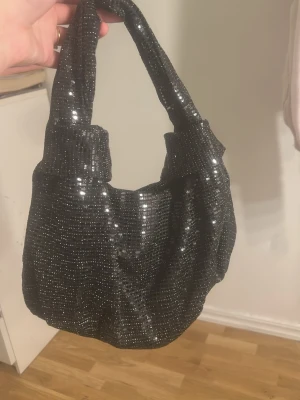 Svart glittrig handväska - Trendig svart handväska med glittriga paljetter som ger en riktigt snygg och festlig look. Väskan har en mjuk, rundad form och ett bredare handtag som gör den bekväm att bära. Perfekt accessoar för att lyfta din outfit. Köpt från Gina tricot aldrig använt