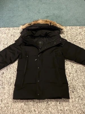 Svart dunjacka Canada Goose XS - Säljer en svart dunjacka från Canada Goose i storlek XS. Jackan har en fluffig pälskant på huvan, diskret logga på ärmen och dragkedja med knappar framtill. Perfekt för kalla dagar och riktigt snygg passform.