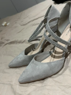 Grå pumps med glittriga band - Snygga grå pumps från WALK med spetsig tå och hög klack. Skorna har två glittriga band över foten som ger en extra touch. Tillverkade i mocka-look för en lyxig känsla och passar perfekt till att lyfta vilken outfit som helst.