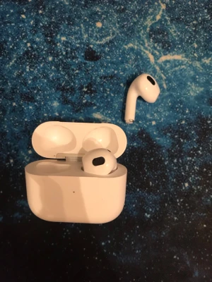 AirPods 3 - Snygga vita trådlösa AirPods från Apple med klassisk stilren design och laddningsetui. Hörlurarna är tillverkade i blank plast och har en smidig form som passar perfekt i öronen. Perfekta för musik, poddar och samtal på språng.