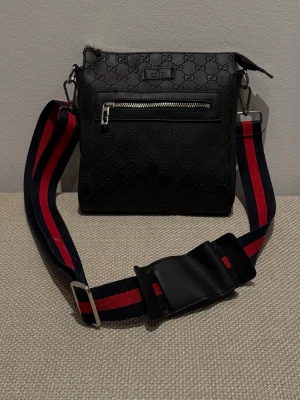 Svart axelväska  - Snygg svart axelväska i skinn. Väskan har en framficka med dragkedja och en bred, justerbar axelrem i marinblått och rött. Perfekt för dig som vill ha en stilren och lyxig accessoar.