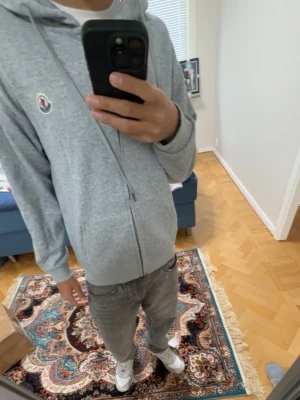 Grå zip hoodie från Moncler - Säljer en stilren grå zip hoodie från Moncler med huva och dragsnören. Hoodien har ett broderat Moncler-märke på bröstet och ribbade muddar vid ärmslut och nederkant. Tillverkad i mjuk bomullsblandning som är skön att bära.