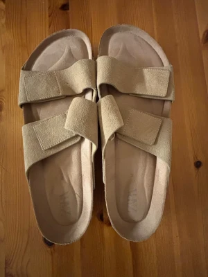 Beige sandaler i mocka från Zara bara använt 2 gånger pga passar inte riktigt - Säljer ett par stilrena beige sandaler från Zara i mjuk mocka. De har två breda remmar med kardborreknäppning och en platt sula för skön komfort. Perfekta för varma dagar och enkel att matcha med olika outfits.