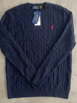 Mörkblå kabelstickad tröja Polo Ralph Lauren - Säljer en mörkblå kabelstickad tröja från Polo Ralph Lauren med klassisk röd logga på bröstet. Tröjan har rund halsringning och långa ärmar. Perfekt till jeans för en clean och stilren look.