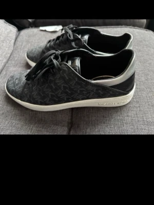 Svarta sneakers från Michael Kors - Snygga svarta sneakers från Michael Kors med silverdetalj på hälen och vit sula. Skorna har ett diskret mönster med MK-logga över hela ovandelen, snörning framtill och är tillverkade i skinn och syntet. Perfekta för dig som vill ha en stilren och trendig look.