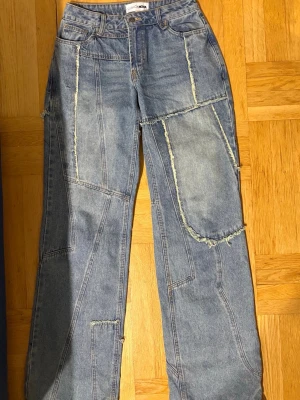 Patchwork jeans från Fashion Nova - Säljer ett par blå patchwork jeans från Fashion Nova med råa kanter och synliga sömmar. Byxorna har en loose passform, hög midja och breda ben. Materialet är klassisk jeans i bomull och de har coola detaljer med fransar längs sömmarna för en edgy vibe. Jeansen har förstört lite vid benet, men det är ganska liten, annars är allt annat super