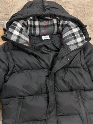 Burberry Jacka - Snygg svart pufferjacka från burburry med huva och rutigt foder i grått, vitt och rött. Jackan har dragkedja framtill, två sidofickor med dragkedja och quiltad design. Perfekt för kalla dagar och ger en cool streetwear-look. Den går även att göra till väst 