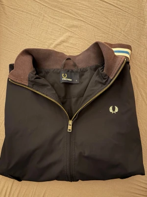 Svart vindjacka från Fred Perry - Snygg svart vindjacka från Fred Perry med guldfärgad dragkedja och klassisk logga på bröstet. Jackan har ribbad brun krage med blå och vita ränder samt en sportig look. Perfekt för dig som vill ha en stilren och tidlös jacka.