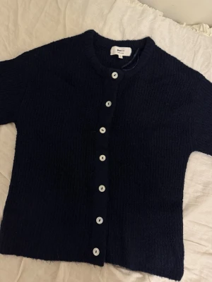 Mörkblå kofta från Gina Tricot - Mysig mörkblå kofta från Gina Tricot i ullblandning. Koftan har rund halsringning, raka ärmar och vita knappar framtill. Perfekt att slänga på sig över en t-shirt eller klänning för en enkel och stilren look.