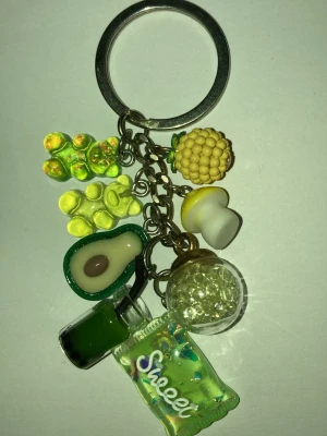 Nyckelring med gröna och gula berlocker - Handgjord nyckelring med flera berlocker i form av olika matvaror som avokado, svamp, vindruvor, dryckesflaska och godispåse. Berlockerna är gjorda i plast och glas och har en lekfull, ungdomlig stil med mycket grönt och gult. Frakt 22-100kr. Kontakta mig om du vill köpa eller har frågor.
