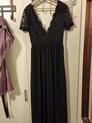 Svart långklänning med spetsdetaljer - Elegant svart långklänning med v-ringning och spets upptill. Klänningen har korta spetsärmar och omlottkänsla vid bysten. Kjoldelen är enkel och faller snyggt. Perfekt för dig som vill ha en klassisk och stilren look.