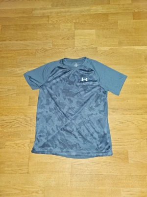 Blå tränings-tshirt från Under Armour - Snygg blå tränings-tshirt från Under Armour med korta ärmar och diskret mönster framtill. Tillverkad i ett lätt och snabbtorkande funktionsmaterial som passar perfekt till gymmet eller löprundan. Logotyp på bröstet och nacken.
