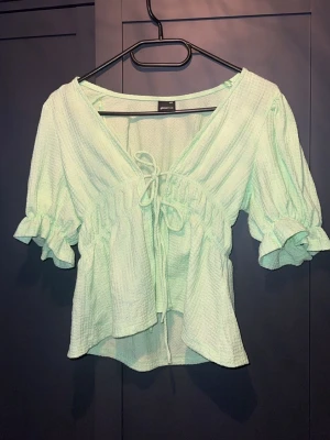 Ljusgrön blus från Gina Tricot - Söt ljusgrön blus från Gina Tricot med v-ringning, knyt framtill och volangdetaljer vid ärmslut.