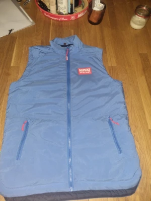 Blå väst från Huski Wear - Blå väst från Huski Wear med dragkedja framtill och två fickor med röda dragkedjor. Västen har hög krage och är tillverkad i ett lätt, vindtätt material. Perfekt för lager-på-lager och outdoor-aktiviteter.ny pris 1000 kr.