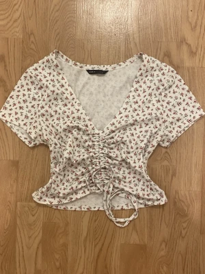 Vit blommig croppad topp med snörning - Supersöt croppad topp med små rosa blommor. Toppen har v-ringning, korta ärmar och snygg snörning framtill som ger en smickrande passform. Perfekt till jeans eller kjol för en trendig look.