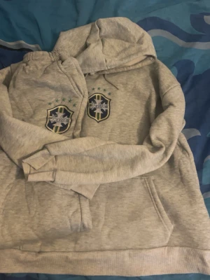Grå brasilien set - Säljer en grå hoodie och byxor i storlek M med Brasilien-inspirerat emblem på bröstet och gröna stjärnor ovanför. Hoodien har huva med dragsko och en stor magficka framtill. Perfekt för dig som gillar sportig stil och vill ha något bekvämt och snyggt. Finns inte många sånna så passa på och köp, skriv för frågor eller prissänkning.