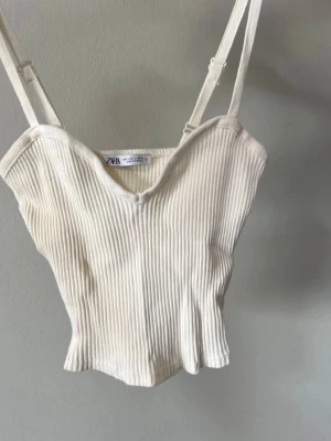 Vit ribbad croppad linnetopp - Säljer en vit croppad linnetopp från Zara med smala justerbara axelband och v-ringad urringning. Toppen är ribbad och har en mjuk, stretchig känsla som gör den superbekväm och enkel att styla till olika looks.