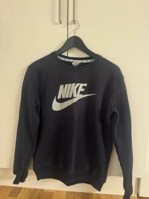 Mörkblå sweatshirt från Nike - Snygg mörkblå sweatshirt från Nike med klassisk vit logga framtill. Tröjan har rund halsringning, ribbade muddar vid ärmslut och nederkant samt en avslappnad passform. Perfekt för en sportig och casual stil.