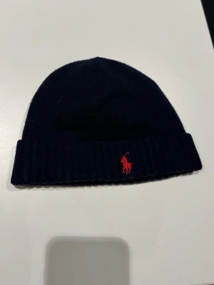 Mörkblå mössa från Polo Ralph Lauren - Snygg mörkblå ribbstickad mössa från Polo Ralph Lauren med klassisk röd logga broderad framtill.  Nästa aldrig använd