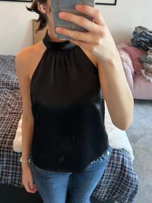 Svart halterneck topp Gina Tricot - Säljer en svart halterneck topp från Gina Tricot storlek 36 i glansigt satinliknande material. Toppen har hög krage med knäppning i nacken och är ärmlös med en lös passform. Perfekt till jeans eller kjol för en snygg look.🖤