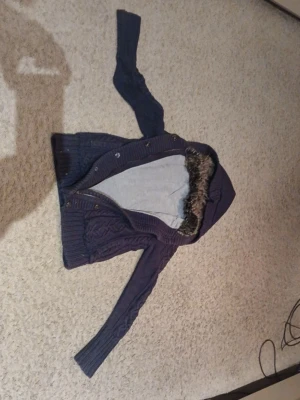 Mörkblå stickad kofta med huva - Mysig mörkblå stickad kofta med huva och fuskpäls runt kanten. Koftan har långa ärmar, knappar framtill och ett snyggt kabelstickat mönster. Perfekt för kyliga dagar när du vill vara både varm och stilren. Skulle säga att den passar Xxs/Xs