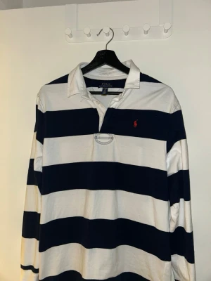 Randig långärmad skjorta Polo Ralph Lauren - Säljer en klassisk långärmad rugbytröja från Polo Ralph Lauren i marinblått och vitt med breda ränder. Skjortan har krage, knappar i halsen och den ikoniska röda loggan broderad på bröstet. Tillverkad i 100% bomull för skön känsla. Skick 7/10 storlek 164 
