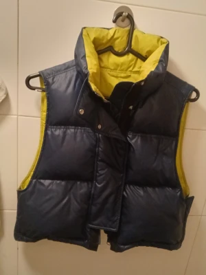 Mörkblå dunväst från ARKET - Snygg mörkblå dunväst från ARKET med kontrasterande gul insida. Västen har hög krage, tryckknappar och dragkedja framtill. Perfekt för lager på lager och ger en sportig vibe. Tillverkad i glansigt material med vadderad känsla.