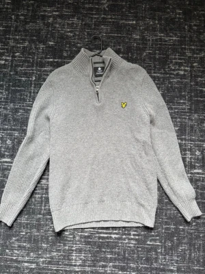 Grå stickad tröja från Lyle & Scott - Grå stickad tröja från Lyle & Scott med halv dragkedja och hög krage. Tröjan har ribbstickade muddar och det klassiska gula logomärket på bröstet. Perfekt för lager-på-lager och snygg till jeans eller chinos.
