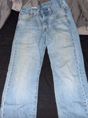 Levi's 501 ljusblå raka jeans - Klassiska ljusblå Levi's 501 jeans med rak passform och fem fickor. Jeansen har knappgylf och den ikoniska läderlappen bak i midjan. Tillverkade i slitstark bomullsdenim, perfekta för en avslappnad och tidlös stil.