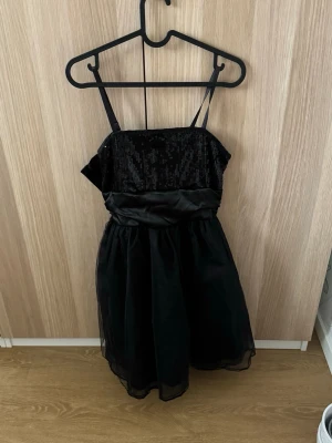 Svart paljettklänning från KappAhl - Snygg svart kortklänning från KappAhl med smala axelband och paljetter upptill. Klänningen har ett glansigt midjeband och ett lätt, transparent tyg över kjolen. Perfekt för dig som vill sticka ut med lite extra glitter. Storleken är för barn men passar mig som har storlek S och även XS.