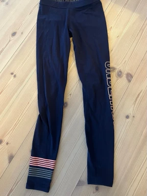 Blå leggings från Under Armour XS - Snygga svarta leggings från Under Armour i storlek XS. De har bred resår i midjan med logga, tryckt text längs benet och coola ränder i rosa, rött och grått vid benslutet. Perfekta för träning eller chill. Materialet är stretchigt och känns riktigt skönt.