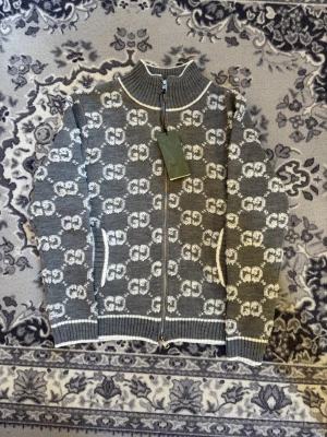 Grå kofta från Gucci med GG-mönster - Säljer en grå och vit kofta från Gucci med ikoniskt GG-mönster över hela plagget. Koftan har ribbad krage, muddar och nederkant, samt dragkedja framtill. Två fickor med vit kant ger extra stil. Perfekt för dig som vill ha något lyxigt och snyggt.