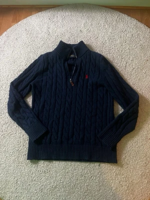 Marinblå stickad tröja Polo Ralph Lauren - Säljer en marinblå stickad tröja från Polo Ralph Lauren med kabelstickat mönster och halv dragkedja vid kragen. Tröjan har långärmad design, ribbade muddar och röd broderad logga på bröstet. Perfekt för dig som gillar klassisk och stilren look.